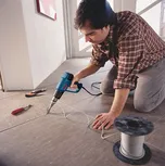 Pessoa ajoelhada a utilizar um soprador térmico para instalar cabos num piso de azulejos.