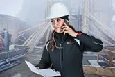 Pessoa que usa equipamento de segurança fala ao telefone e segura um caderno num estaleiro de obras.