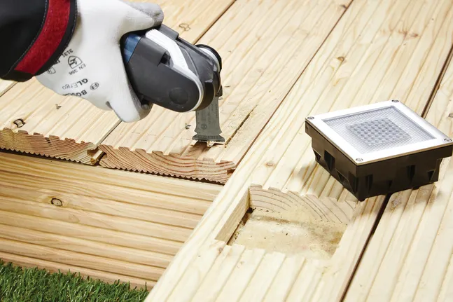 Uma pessoa que utilize equipamento de segurança utiliza uma ferramenta elétrica para cortar um painel de madeira de um deck.