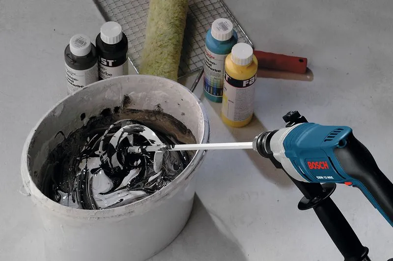 Misturador elétrico misturando tinta num balde ao lado de vários frascos de tinta e um rolo.