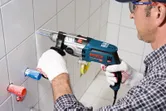Uma pessoa que utilize equipamento de segurança fura uma parede de azulejos com um berbequim de impacto.