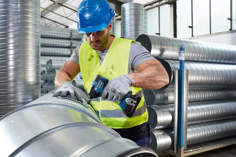 Uma pessoa que utiliza equipamento de segurança fixa uma tubagem metálica utilizando uma ferramenta elétrica sem fios.