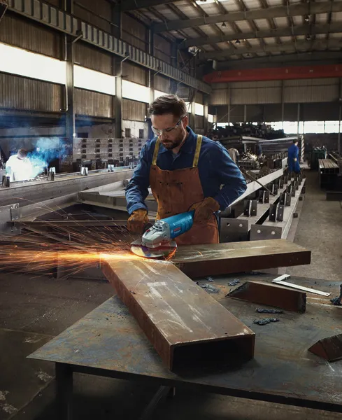 Uma pessoa que usa equipamento de segurança retifica uma grande viga metálica numa oficina industrial.