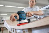 Uma pessoa que utiliza equipamento de segurança lixa uma superfície curva de madeira com uma ferramenta elétrica.