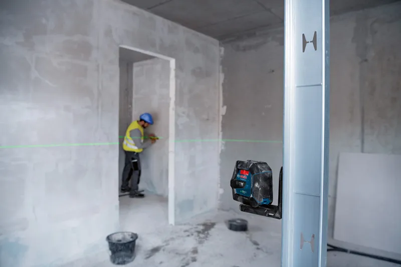 Uma pessoa que use equipamento de segurança marca uma parede guiada por uma ferramenta de nivelamento a laser verde.