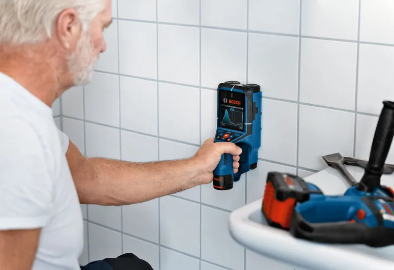 Uma pessoa examina uma parede de azulejos com uma ferramenta de inspeção num espaço de trabalho.