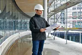 Pessoa que usa equipamento de segurança e casaco aquecido analisa plantas num estaleiro de obras.
