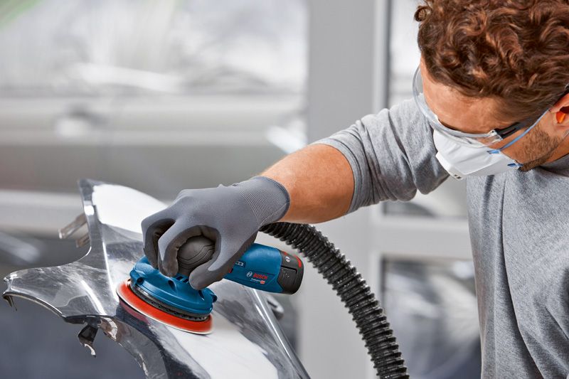 Uma pessoa que utiliza equipamento de segurança lixa uma peça de automóvel com uma lixadora orbital sem fios.