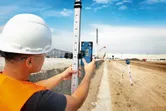 Uma pessoa que usa equipamento de segurança alinha um recetor laser numa mira de nivelamento num estaleiro de obras.