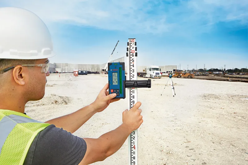 Uma pessoa que utilize equipamento de segurança utiliza um recetor laser numa mira de medição num estaleiro de obras.