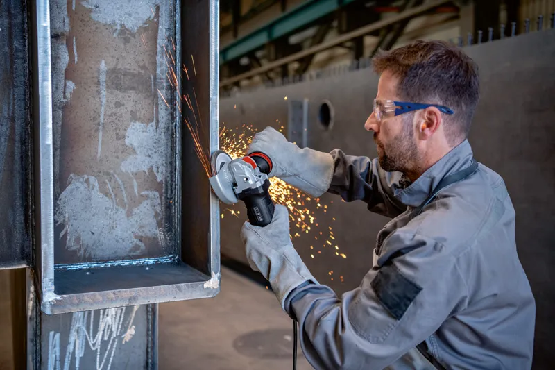 Uma pessoa que usa equipamento de segurança tritura uma viga de metal com uma rebarbadora, e as faíscas voam.