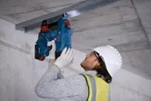 Uma pessoa que utiliza equipamento de segurança fixa um canal de metal a um teto de betão com um pregador de betão sem fios.