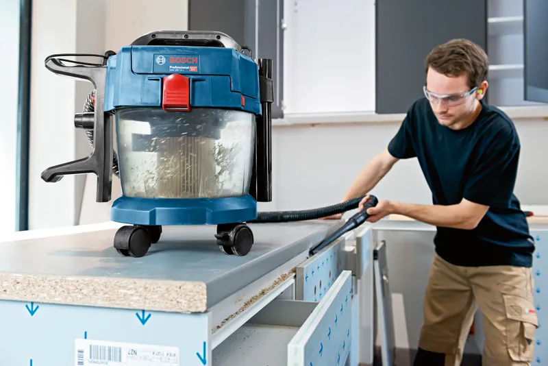 Uma pessoa que utiliza equipamento de segurança utiliza um aspirador de pó e água para limpar uma bancada de trabalho.