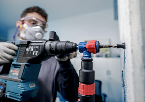 Uma pessoa que utilize equipamento de segurança fura uma parede utilizando um berbequim de impacto.