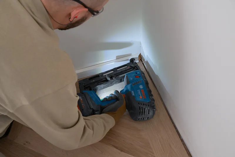 Uma pessoa que utiliza equipamento de segurança utiliza um prego de madeira sem fios para fixar o acabamento ao chão.