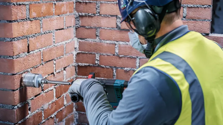 Uma pessoa que utiliza equipamento de segurança perfura uma parede de tijolos com uma ferramenta elétrica.