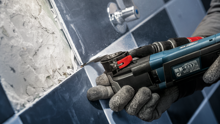 Uma pessoa que utiliza equipamento de segurança remove o betume de azulejos utilizando uma ferramenta multifuncional elétrica.