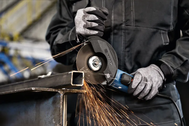 Uma pessoa que utiliza equipamento de segurança tritura uma viga de metal com uma rebarbadora, criando faíscas.