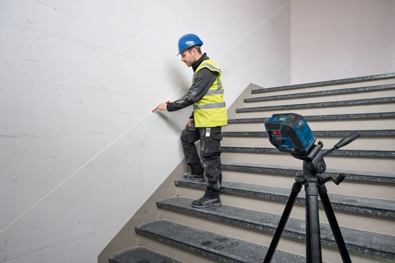 Uma pessoa que utilize equipamento de segurança marca uma parede na escada utilizando uma ferramenta de nivelamento a laser.