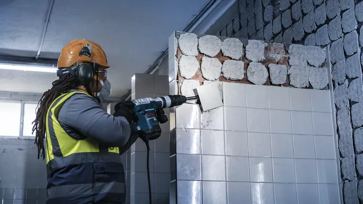 Uma pessoa que utiliza equipamento de segurança remove azulejos da parede com uma ferramenta elétrica.