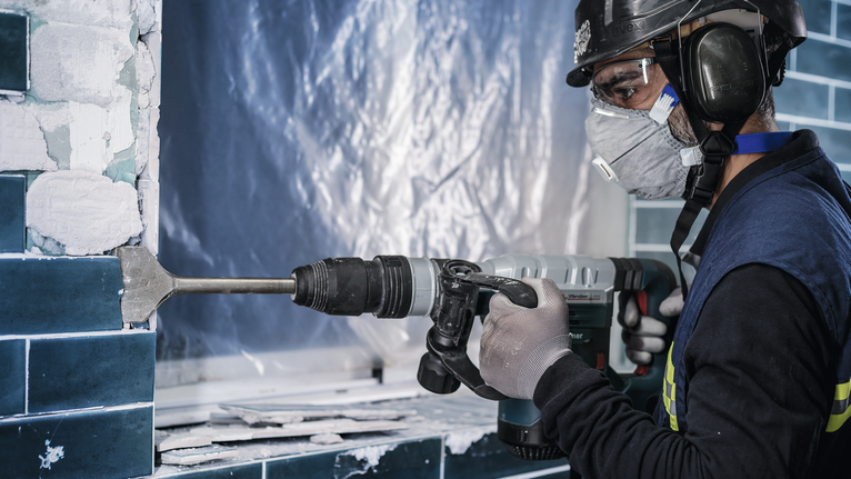 Uma pessoa que utiliza equipamento de segurança remove azulejos da parede utilizando um martelo rotativo.