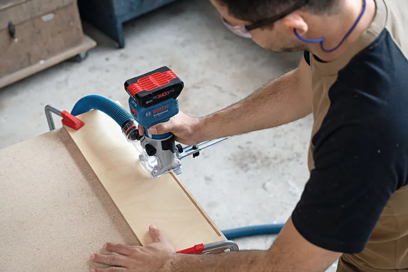 Uma pessoa que utiliza equipamento de segurança utiliza uma fresadora sem fios para moldar madeira numa bancada de trabalho.
