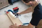 Uma pessoa que utiliza equipamento de segurança utiliza uma fresadora sem fios para moldar madeira numa bancada de trabalho.