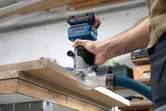 Uma pessoa utiliza um router portátil com extração de pó na borda de uma placa de madeira.