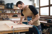 Uma pessoa que usa equipamento de segurança utiliza uma fresadora de madeira numa tábua fixada numa oficina.