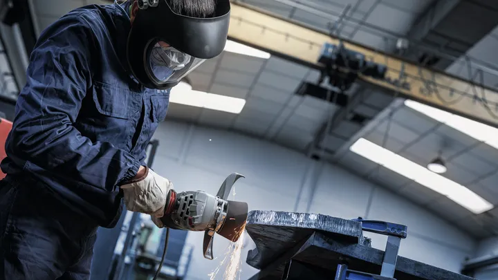 Uma pessoa que utilize equipamento de segurança retifica uma viga metálica numa bancada de trabalho.