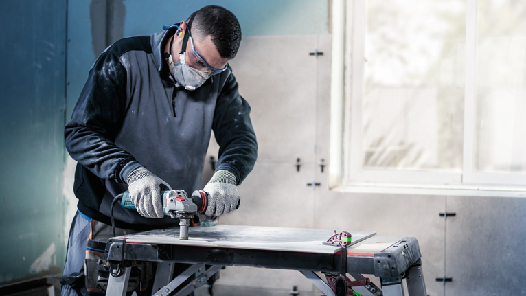 Pessoa que utiliza equipamento de segurança a cortar azulejos com uma ferramenta elétrica numa bancada de trabalho.