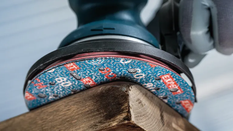 Pessoa que utiliza equipamento de segurança a lixar o canto de uma tábua de madeira.