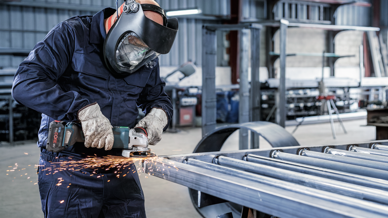 Pessoa que utiliza equipamento de segurança retifica vigas metálicas numa oficina industrial.