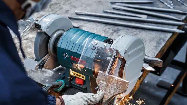 Um trabalhador que utilize equipamento de segurança esmerilhe uma peça de metal com uma rebarbadora de bancada.