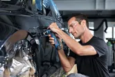 Uma pessoa que utilize equipamento de segurança lixa a porta de um automóvel com uma lixadora orbital sem fios.