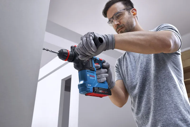 Uma pessoa que utilize equipamento de segurança perfura uma parede de betão utilizando um martelo rotativo sem fios.