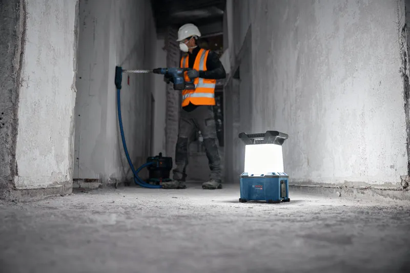 Um trabalhador que usa equipamento de segurança perfura uma parede e uma luz de trabalho sem fios está no chão.