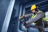 Uma pessoa que utiliza equipamento de segurança utiliza uma rebarbadora angular para cortar metal dentro de uma estrutura grande.