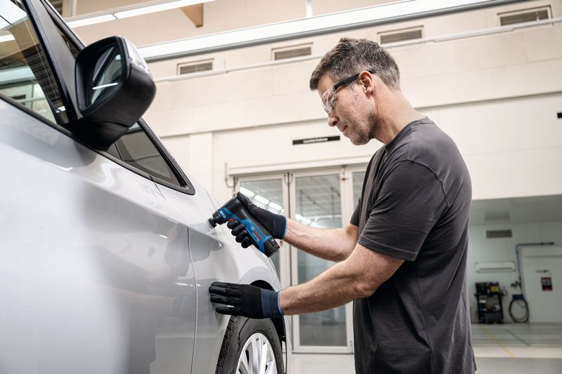 Uma pessoa que utiliza equipamento de segurança lixa a porta de um automóvel utilizando uma lixadora orbital.