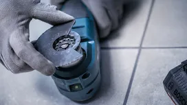 Pessoa que utiliza equipamento de segurança fixa uma lâmina circular a uma ferramenta elétrica num piso de azulejos.