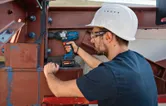 Uma pessoa que utilize equipamento de segurança utiliza uma chave de impacto sem fios numa viga de aço.