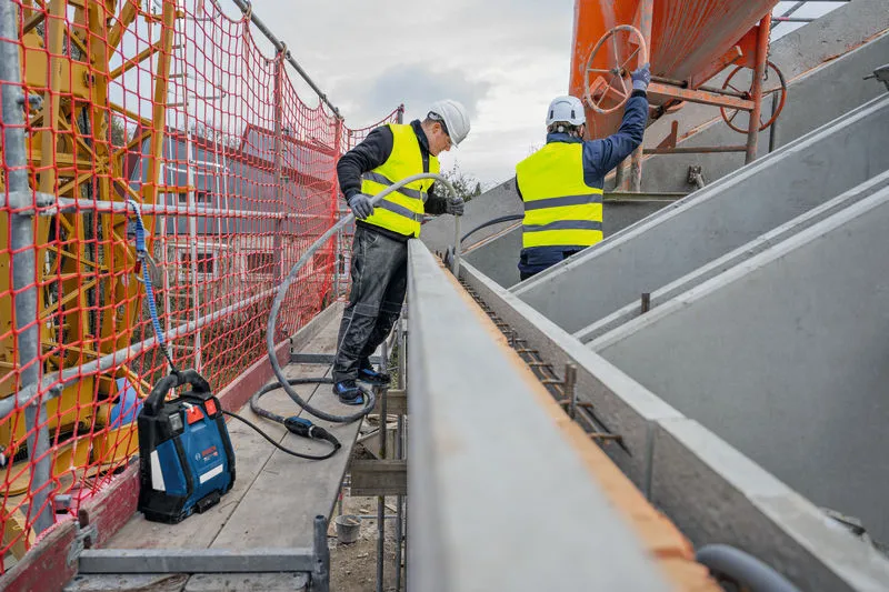 Duas pessoas que usam equipamento de segurança trabalham num andaime com ferramentas de construção e formas de betão.