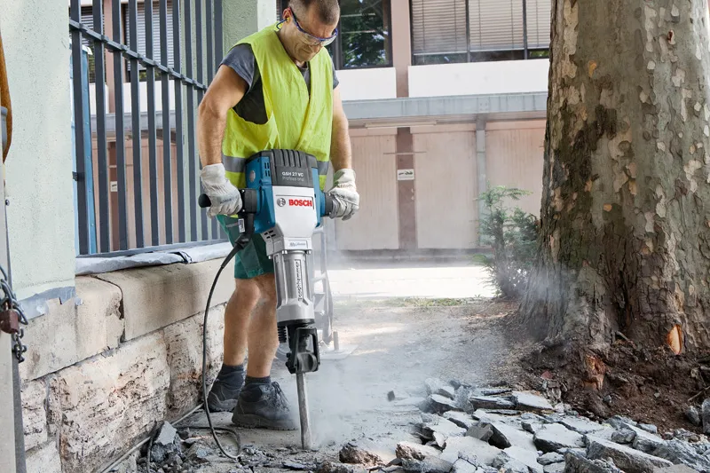 Uma pessoa que utiliza equipamento de segurança quebra o pavimento ao ar livre com um disjuntor elétrico de grandes dimensões.