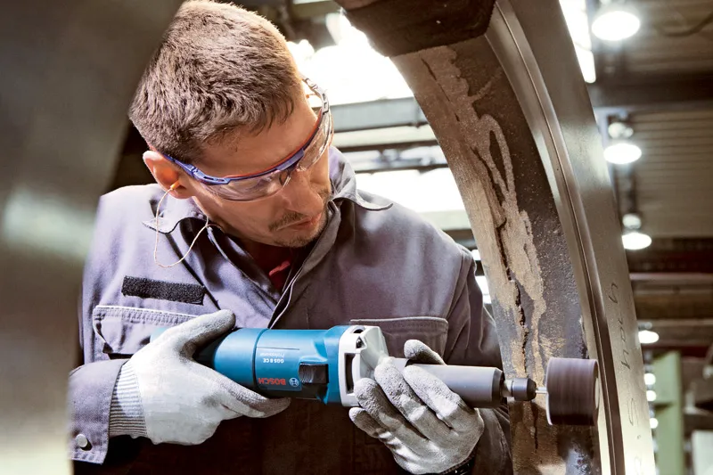 Uma pessoa que utiliza equipamento de segurança esmerilha a superfície interior de um grande anel de metal com uma rebarbadora reta.