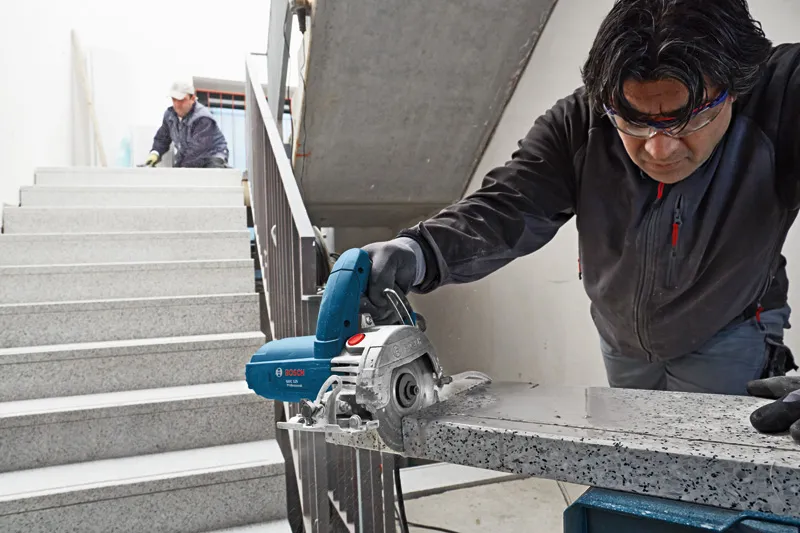 Una persona que usa equipo de seguridad utiliza un cortador de baldosas de diamante para cortar una losa de piedra.
