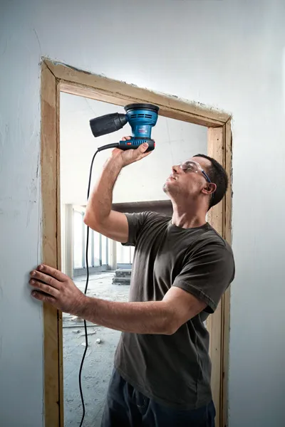 Una persona que usa equipo de seguridad lija un marco de puerta de madera con una lijadora eléctrica.