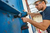 Una persona que usa equipo de seguridad utiliza una llave de impacto inalámbrica sobre una viga de metal de gran tamaño.