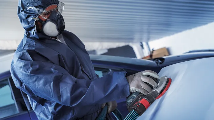 Una persona que usa equipo de seguridad lija la superficie de un automóvil con una lijadora eléctrica.
