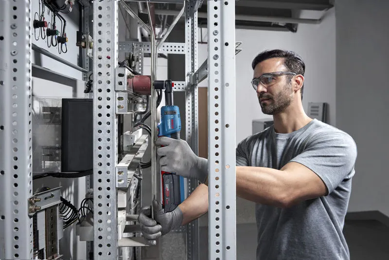 Una persona que usa equipo de seguridad utiliza una llave de trinquete inalámbrica en un estante de metal.