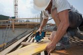 Un trabajador que usa equipo de seguridad corta una tabla de madera con una sierra caladora inalámbrica en un sitio de construcción.